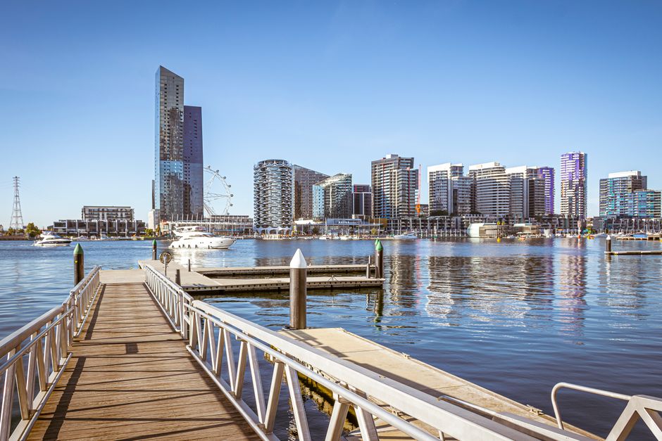 A wooden dock extending over calm waters leads towards a modern city skyline featuring high-rise buildings with varied architectural styles, including glass facades reflecting the blue sky. Several boats are moored along the waterfront, with some positioned near the dock and others further out on the water. The dock has metal railings painted white with green-tipped posts, and a cleat visible on the deck for securing vessels. In the background, contemporary buildings of different heights and designs dominate the skyline, some with curved or textured exteriors, and a Ferris wheel partially visible behind the structures. The scene is illuminated by natural daylight, casting minimal shadows and creating a clear, bright atmosphere. This setting illustrates an urban waterfront environment that could relate to private or independent waste management solutions, such as rubbish collection services, situated in a lively metropolitan area with marina amenities, aligning subtly with the topic of alternative rubbish removal options like those provided by Waste Collection Docklands.