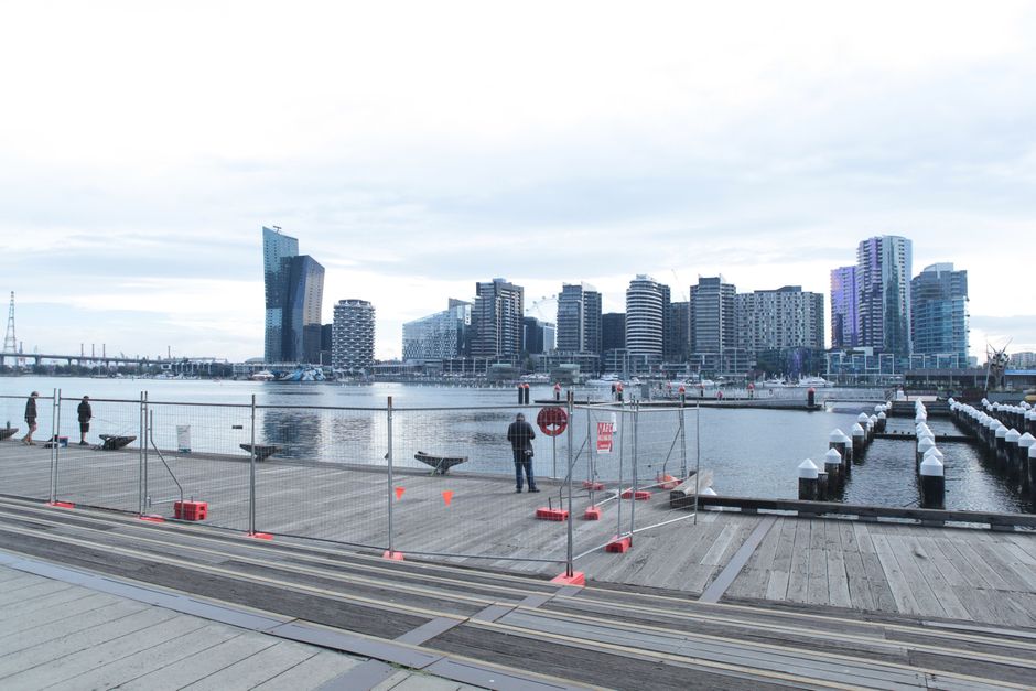 A view of a waterfront urban area with a wooden dock extending over calm water, enclosed by temporary metal fencing and safety barriers with orange cones and red signs. Several people are standing along the dock, some near the fencing, and one is operating or inspecting equipment. In the background, a modern city skyline features high-rise buildings with glass facades, including a distinctive angular skyscraper, under a cloudy sky. The scene suggests an area undergoing construction, maintenance, or other temporary work related to the waterfront environment, highlighting the potential for professional waste or debris management during site activities, as provided by waste collection services in docklands areas.