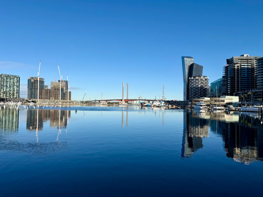 A wooden dock extending over calm waters leads towards a modern city skyline featuring high-rise buildings with varied architectural styles, including glass facades reflecting the blue sky. Several boats are moored along the waterfront, with some positioned near the dock and others further out on the water. The dock has metal railings painted white with green-tipped posts, and a cleat visible on the deck for securing vessels. In the background, contemporary buildings of different heights and designs dominate the skyline, some with curved or textured exteriors, and a Ferris wheel partially visible behind the structures. The scene is illuminated by natural daylight, casting minimal shadows and creating a clear, bright atmosphere. This setting illustrates an urban waterfront environment that could relate to private or independent waste management solutions, such as rubbish collection services, situated in a lively metropolitan area with marina amenities, aligning subtly with the topic of alternative rubbish removal options like those provided by Waste Collection Docklands.
