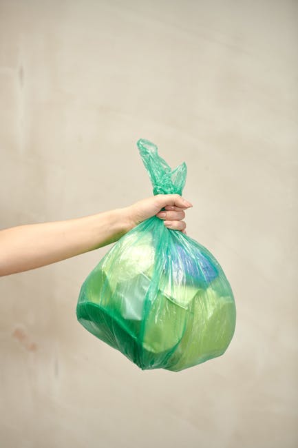 A person's arm and hand are visible, holding a green plastic rubbish bag tightly by its twisted knot at the top. The bag appears to be filled with waste, causing it to bulge and take on a rounded shape. The plastic is semi-transparent, allowing some of the contents to be faintly seen inside, and the surface has a slightly crinkled texture reflecting light. The background is a plain, neutral-toned wall, providing no additional environmental context. The scene is lit with soft, diffuse lighting that highlights the subtle creases and contours of the bag, emphasizing its volume and the act of rubbish collection. The image focuses on an individual’s effort in waste disposal, illustrating private rubbish handling, which may relate to independent waste collection or onsite clearance services provided by waste management companies like Waste Collection Docklands. This simple, clear depiction emphasizes the physical aspect of rubbish removal through flexible, environmentally conscious waste disposal practices.