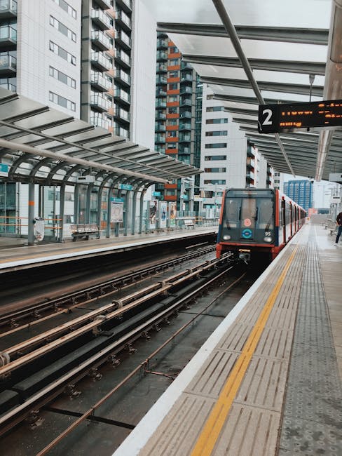 A modern urban train station platform with a light grey tactile paving strip along the edge, and a red and black train approaching from the right side of the image. The train has a sleek, smooth finish with large front windows and visible doors on the side. Above the platform, there's a glass and metal canopy supported by black metal beams, providing shelter for waiting passengers. Adjacent to the platform, an overhead electronic sign displays platform information with illuminated orange digital characters. In the background, tall high-rise buildings with various glass and concrete facades are visible, creating a dense cityscape setting. The environment is illuminated by natural daylight, with a slightly overcast sky, adding a neutral, professional atmosphere typical of busy city transport hubs where efficient passenger movement and potential for alternative waste management strategies, like private collection or on-site clearance, are relevant considerations for operators such as Waste Collection Docklands.