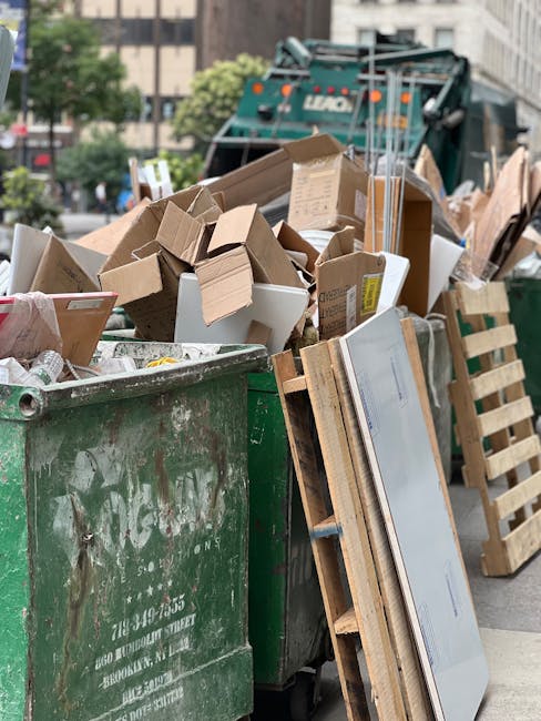 A high-angle view of a cluttered outdoor area showing a large pile of mixed waste materials including plastic bags, cardboard boxes, and discarded household items. In the foreground, a white waste collection truck with black lettering and registration number AZ 99-FX-185 is parked on uneven, gravelly ground. Three individuals, wearing casual clothing and work gloves, are actively engaged in clearing and lifting debris from the ground. The area is surrounded by various discarded objects such as wooden spools, metal pipes, and broken appliances, indicating an informal waste dumping site or site for waste removal. The environment appears cluttered and unmanaged, with environmental context suggesting the need for professional rubbish removal services, such as those provided by Waste Collection Docklands, to handle private or alternative waste disposal options in urban or temporary outdoor settings.