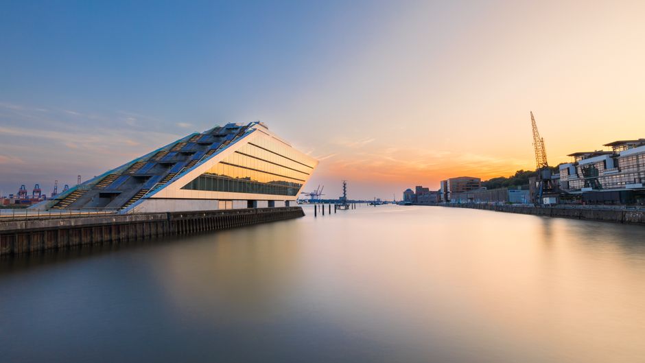 A wide-angle view of the Docklands waterfront during sunset, featuring a modern, angular building with a distinctive sloped roof on the left side, its exterior clad in reflective glass and metallic panels that catch the warm tones of the sky. The building is situated on a concrete quay with a textured surface, extending into calm water that reflects the soft orange and pink hues of the evening sky. In the background, cargo cranes and industrial infrastructure are visible along the horizon, framing the scene with a mixture of shipping and dockside elements. To the right, a cluster of contemporary buildings, with glass facades and angular shapes, line the waterfront, complemented by a tall crane and other port equipment. The overall environment suggests an active dockside area, where private waste collection services like Waste Collection Docklands may operate, particularly for event-related rubbish removal, with the serene water and clear evening atmosphere highlighting the urban landscape and industrial port activity.