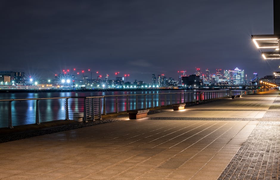 A wide-angle view of the Docklands waterfront during sunset, featuring a modern, angular building with a distinctive sloped roof on the left side, its exterior clad in reflective glass and metallic panels that catch the warm tones of the sky. The building is situated on a concrete quay with a textured surface, extending into calm water that reflects the soft orange and pink hues of the evening sky. In the background, cargo cranes and industrial infrastructure are visible along the horizon, framing the scene with a mixture of shipping and dockside elements. To the right, a cluster of contemporary buildings, with glass facades and angular shapes, line the waterfront, complemented by a tall crane and other port equipment. The overall environment suggests an active dockside area, where private waste collection services like Waste Collection Docklands may operate, particularly for event-related rubbish removal, with the serene water and clear evening atmosphere highlighting the urban landscape and industrial port activity.